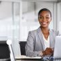 Portrait of a cheerful businesswoman sitting at desk in modern office and looking at camera. Attractive and successful black business woman sitting at conference table with laptop. Smiling african american executive using laptop while working from office with copy space.